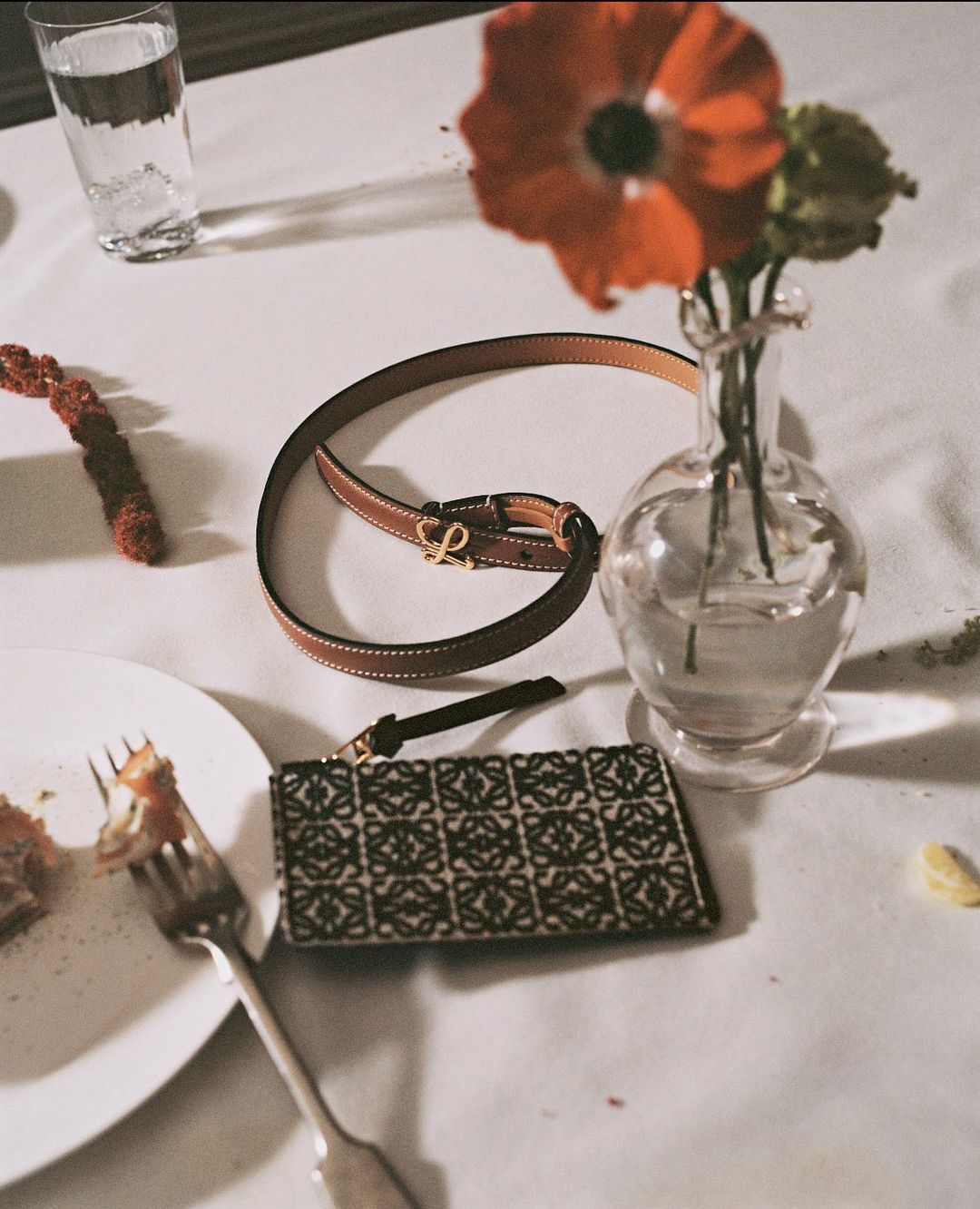 Still life of a brown leather belt, patterned wallet, water glass, orange flower in vase, and half-eaten plate on a white table.