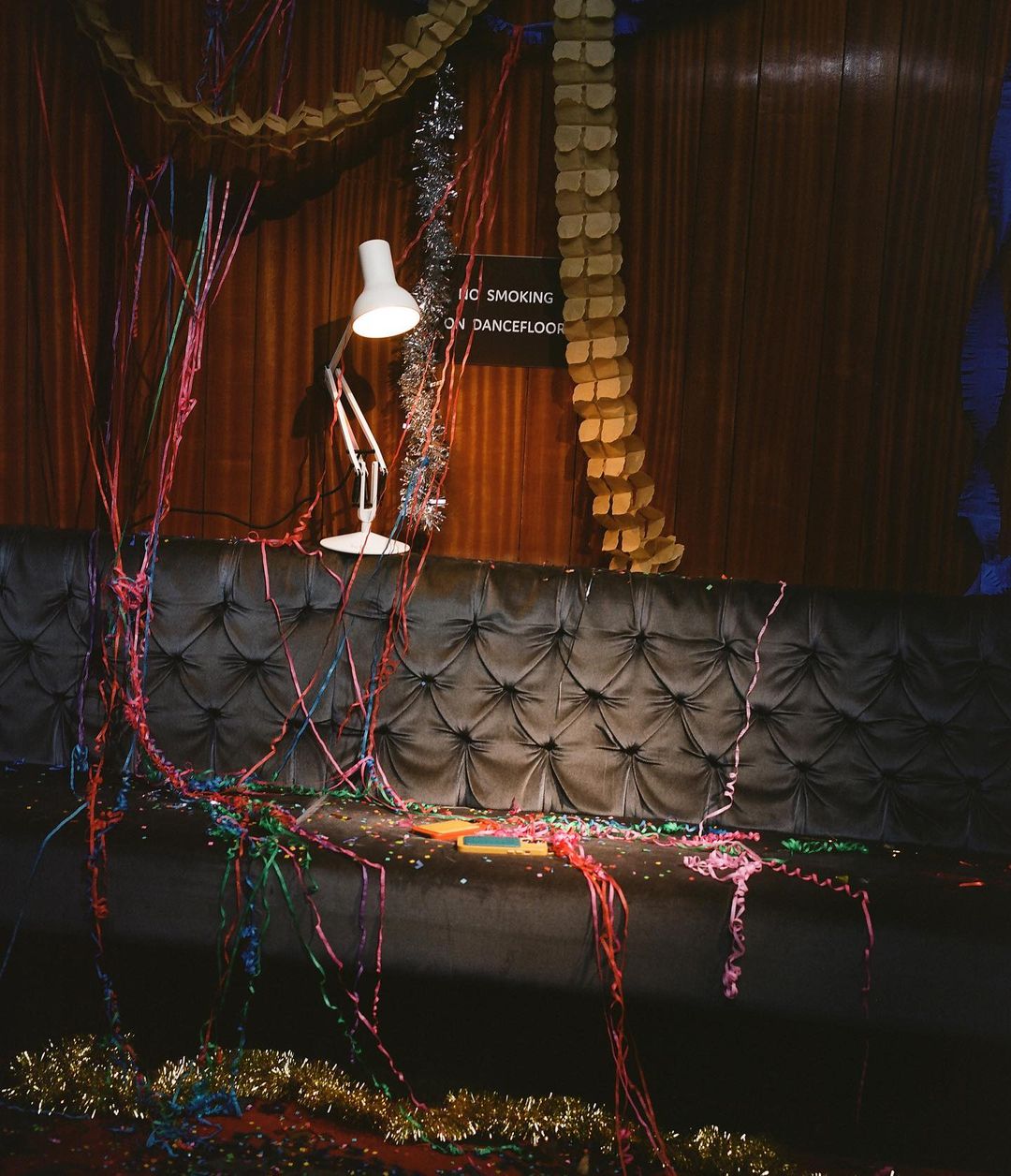 Celebratory scene with streamers and confetti scattered around a black tufted couch and a white lamp in an indoor setting.