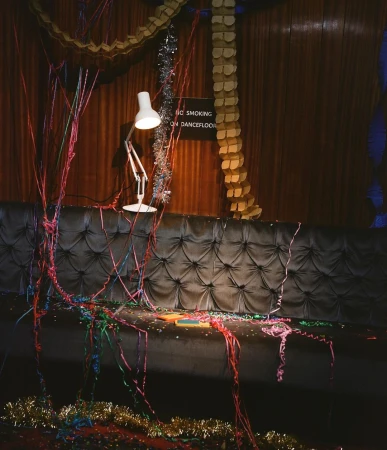 Celebratory scene with streamers and confetti scattered around a black tufted couch and a white lamp in an indoor setting.