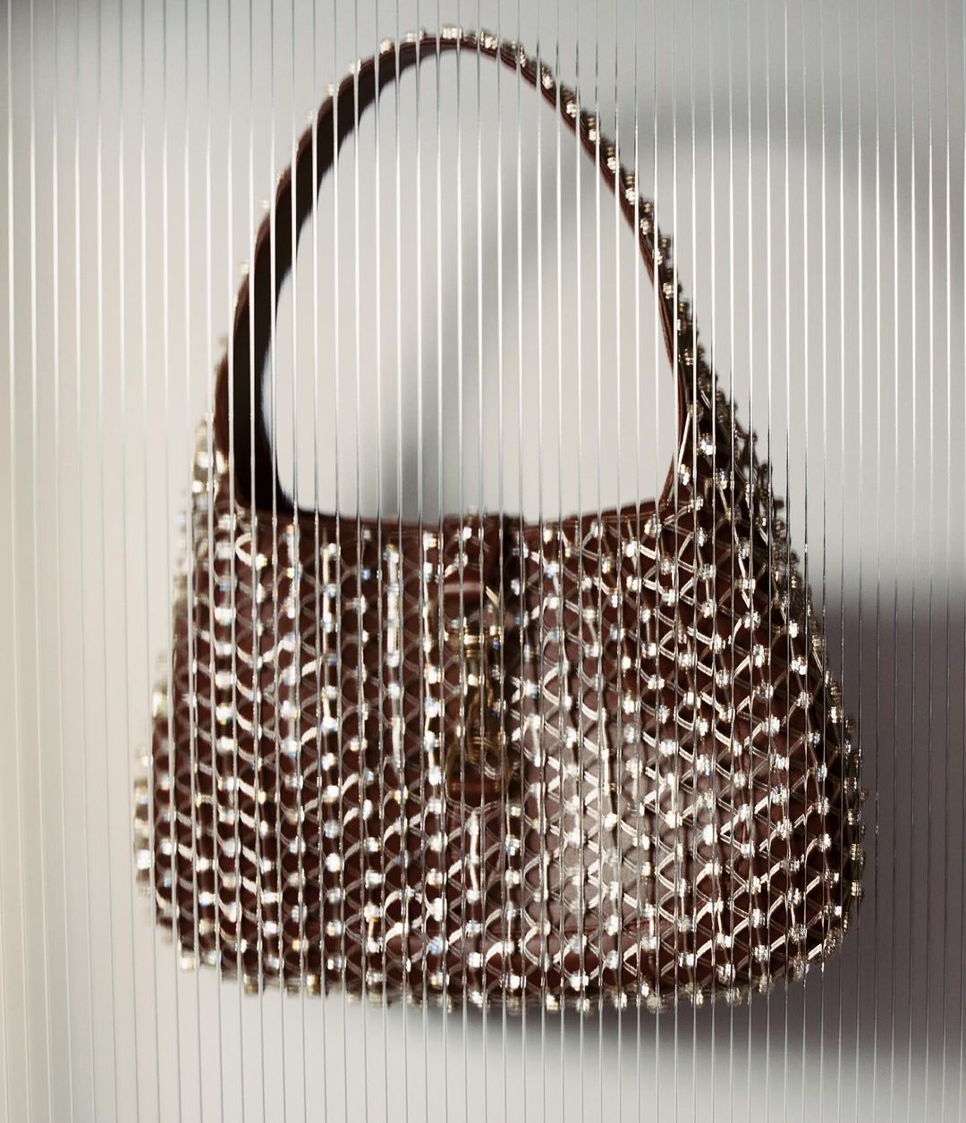 Close-up studio shot of a brown hobo-style handbag adorned with silver-toned studs, against a vertically striped gray and white backdrop.