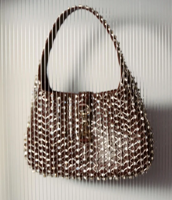 Close-up studio shot of a brown hobo-style handbag adorned with silver-toned studs, against a vertically striped gray and white backdrop.
