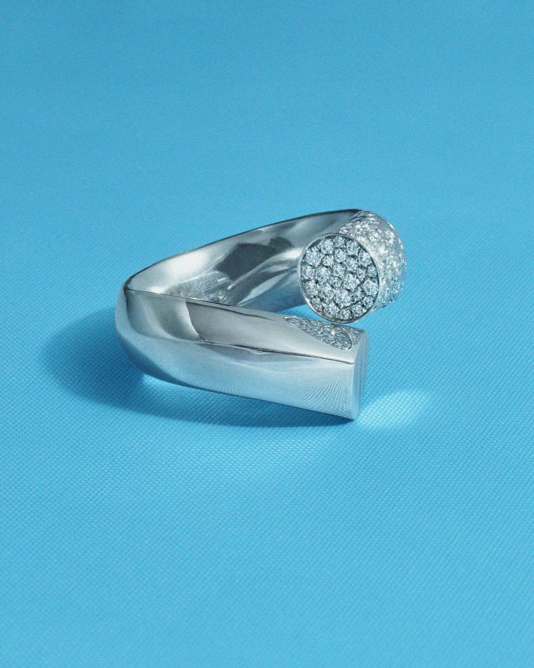 Close-up of a modern silver metal ring set with sparkling diamonds on a smooth blue surface in a jewelry or fashion context.