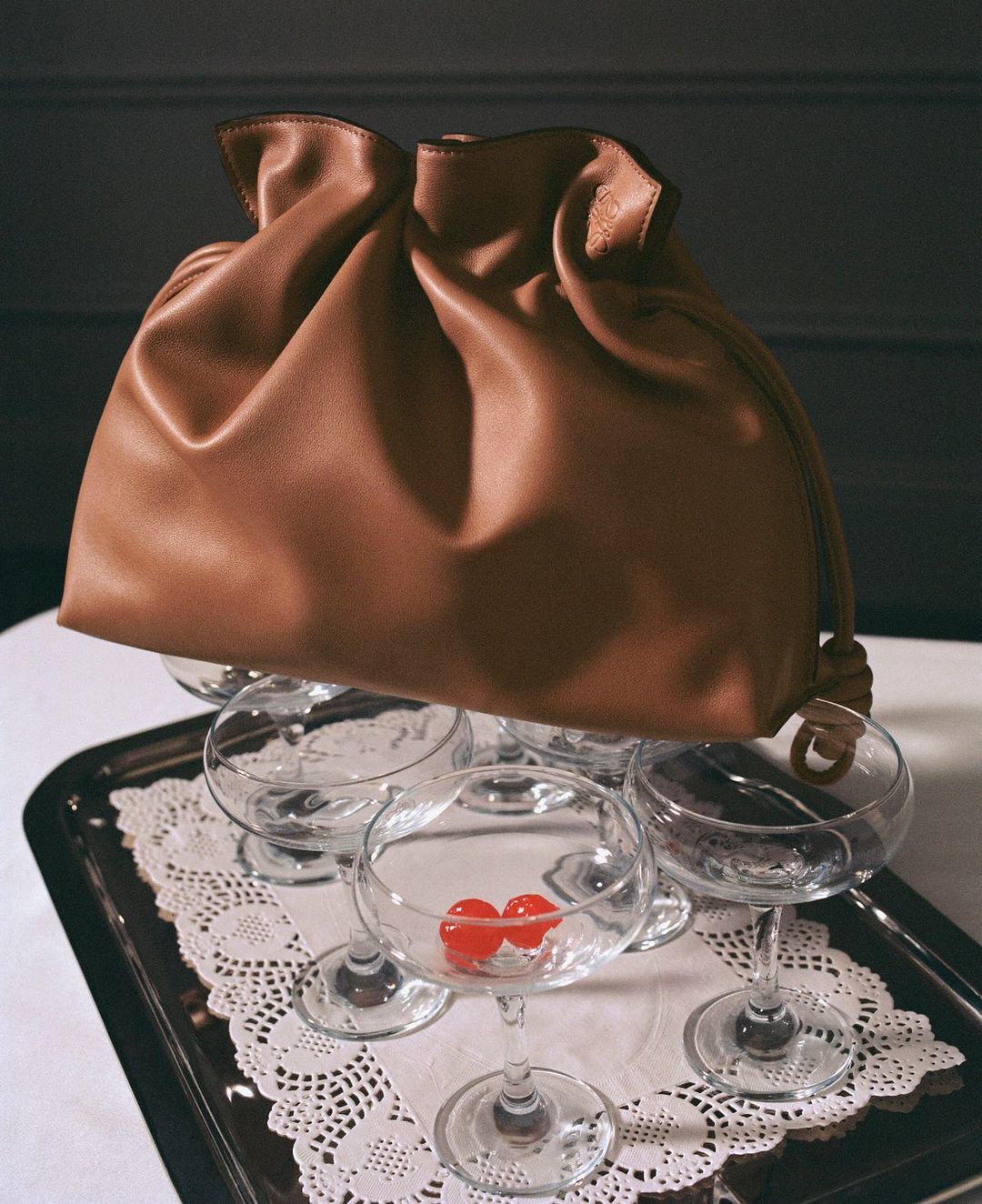Still life shot of a brown leather drawstring bag balancing atop stemmed cocktail glasses, with cherries inside, on a silver tray.