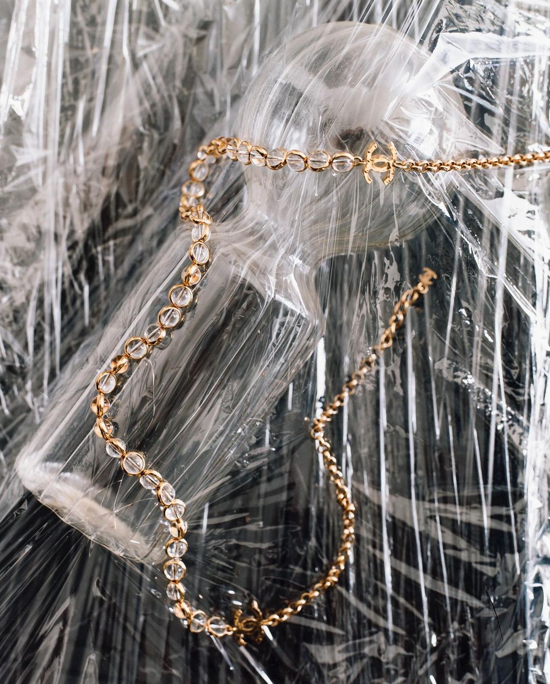 Gold necklace with crystal beads and logo charms displayed over wrinkled plastic wrapping. Jewellery flat lay for fashion or beauty.
