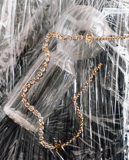 Gold necklace with crystal beads and logo charms displayed over wrinkled plastic wrapping. Jewellery flat lay for fashion or beauty.