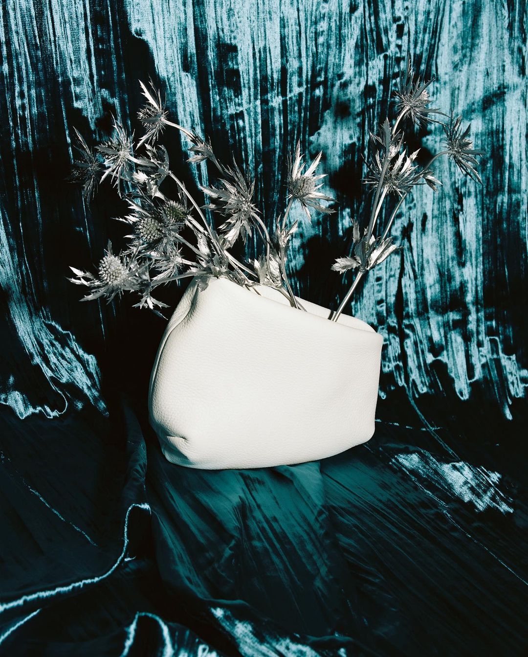 Still life studio shot of a white, leather vase holding thistle flowers, set against a textured turquoise and black background.