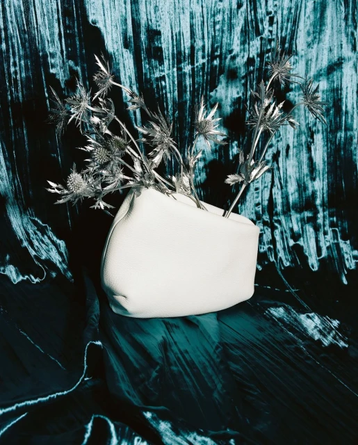 Still life studio shot of a white, leather vase holding thistle flowers, set against a textured turquoise and black background.