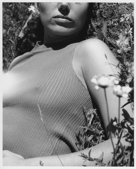 Monochrome portrait of a woman lying in grass with white flowers, wearing a ribbed top. Upper body is visible in a close-up shot.