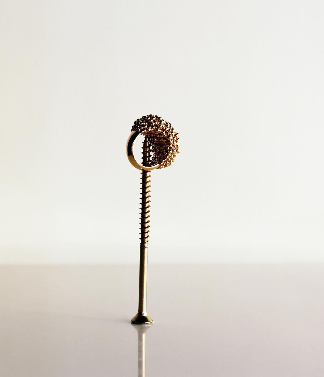 Close-up studio shot of a gold metal ring with textured design atop a gold-colored screw standing upright on a reflective surface.