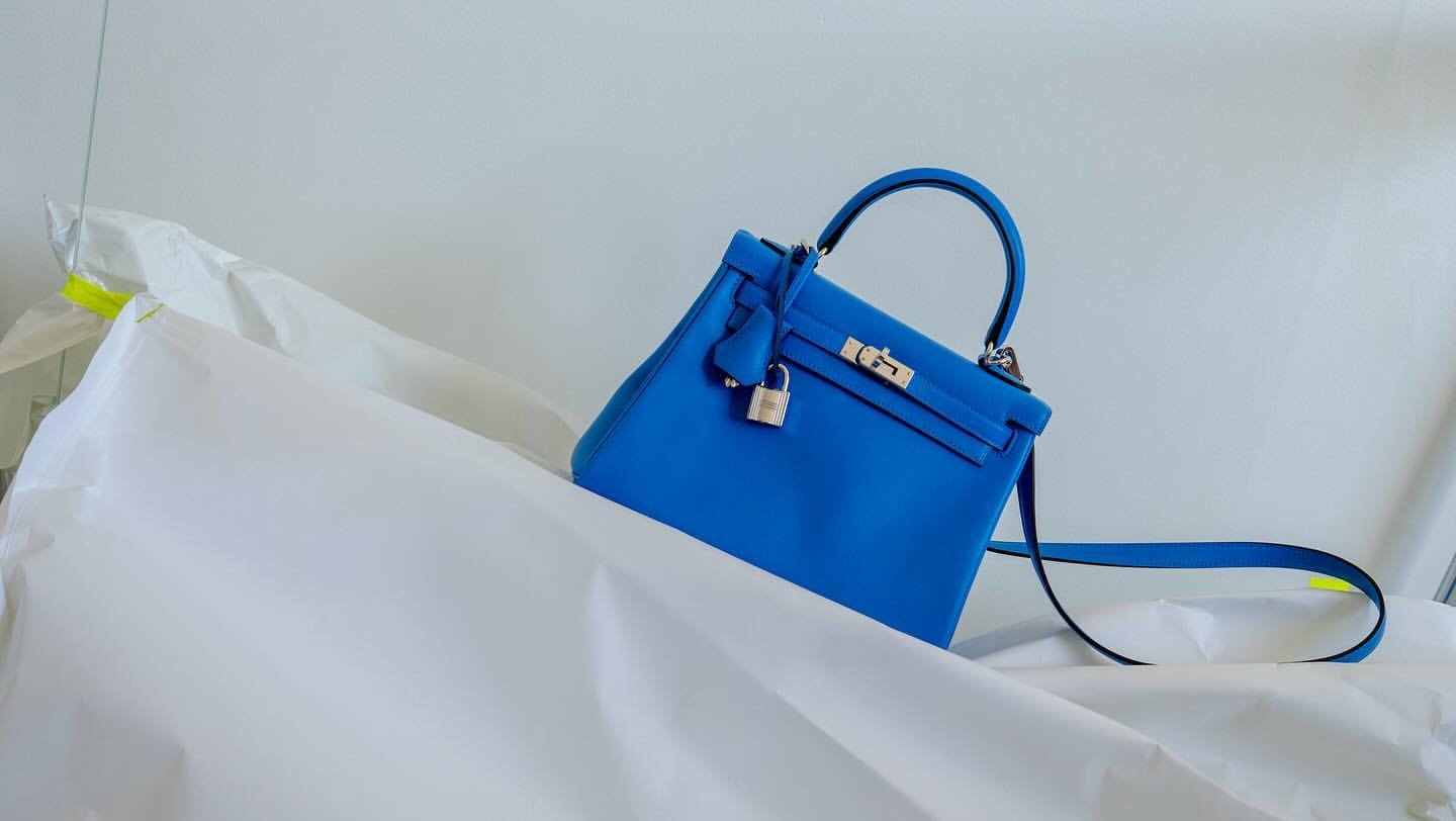 Bright blue Kelly bag by Hermès on draped white fabric. Luxury handbag with top handle, lock, and silver hardware is the focal point.