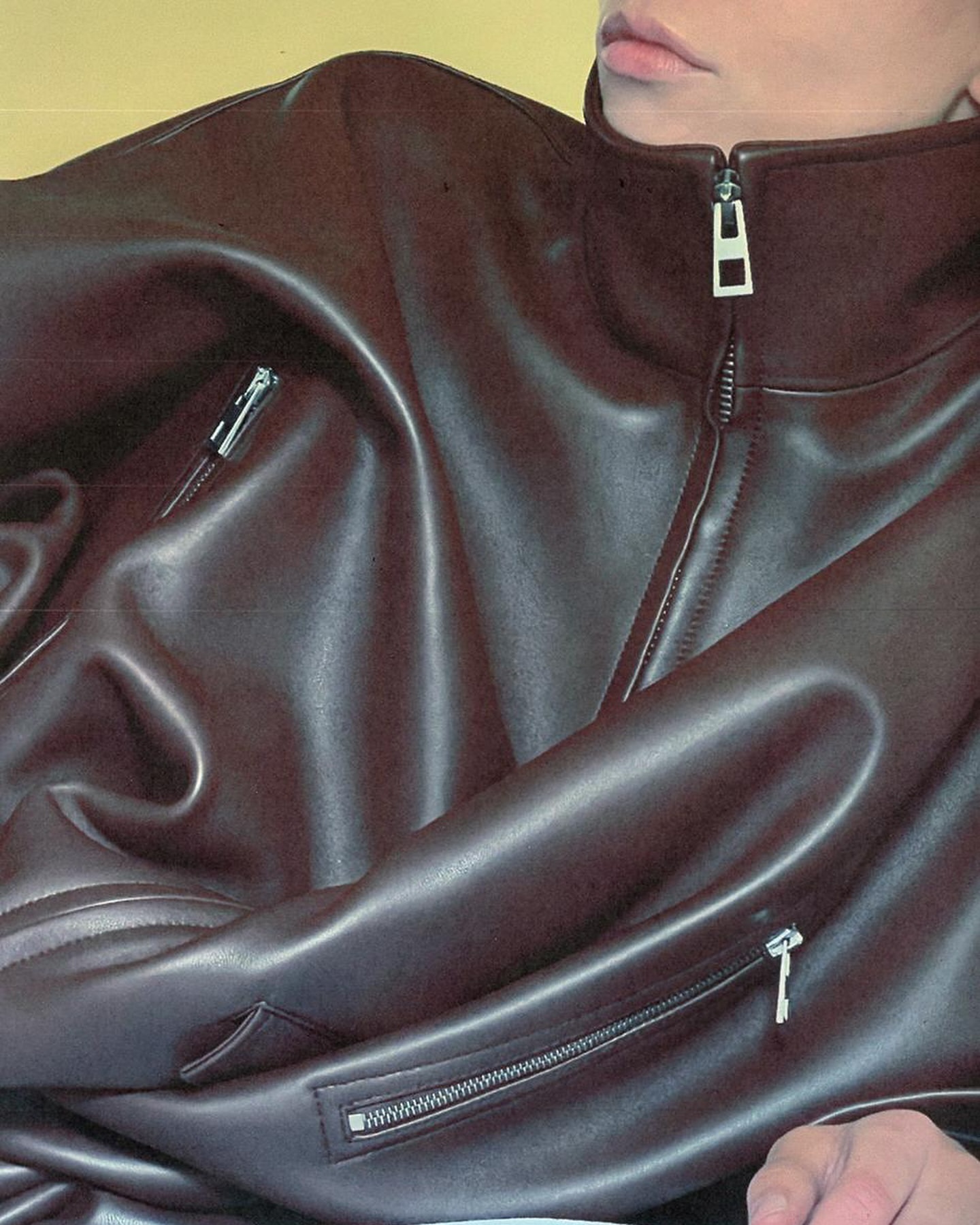 Close-up of a person wearing a shiny, dark maroon faux leather zip-up jacket with silver hardware, set against a light yellow backdrop.