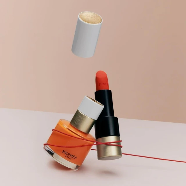 Still life featuring floating cosmetics: lipstick, nail polish, and a cylindrical container, all connected with a thin red string on a light pink backdrop.