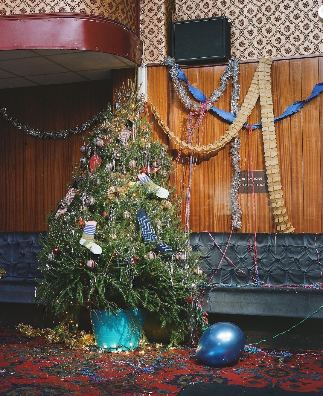 Decorated Christmas tree with ornaments and socks in a vintage interior with patterned wallpaper, wood paneling, and party decorations.