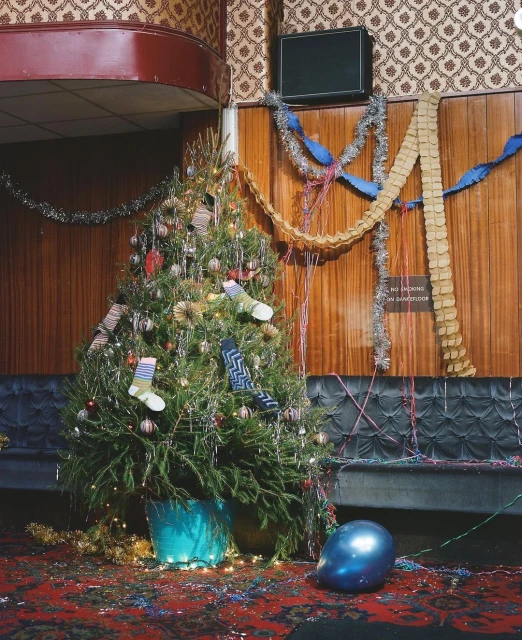 Decorated Christmas tree with ornaments and socks in a vintage interior with patterned wallpaper, wood paneling, and party decorations.