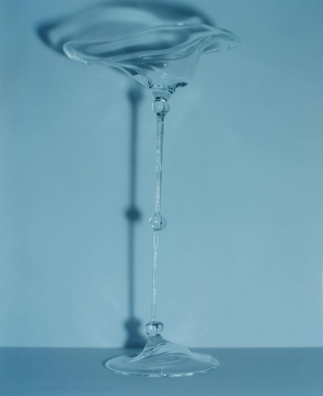 Conceptual studio photograph of an upside-down, clear glass goblet, with its shadow visible against a solid, light blue background.