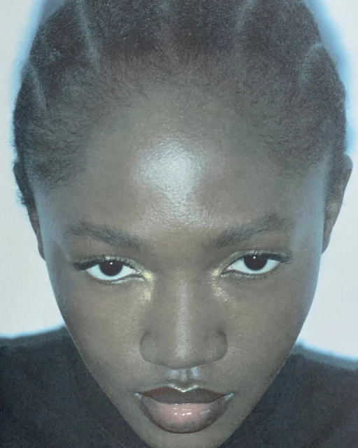 Close-up portrait of an adult female with dark skin, glossy lips, and braided hair, facing the camera with a neutral facial expression.