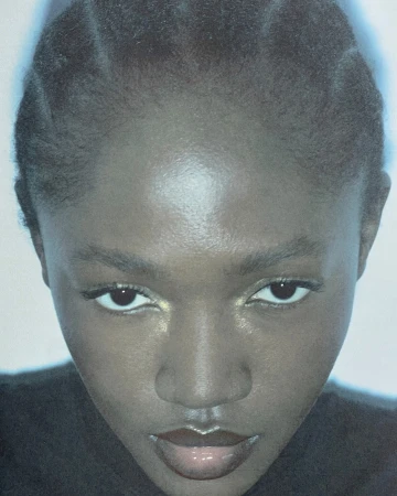 Close-up portrait of an adult female with dark skin, glossy lips, and braided hair, facing the camera with a neutral facial expression.