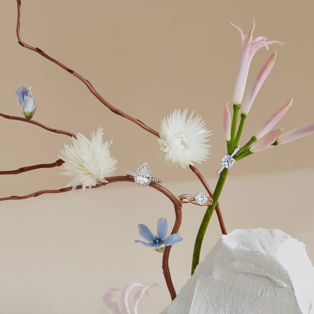 Close-up of four diamond engagement rings displayed on branches and flowers, featuring various cuts like pear, round, and princess.