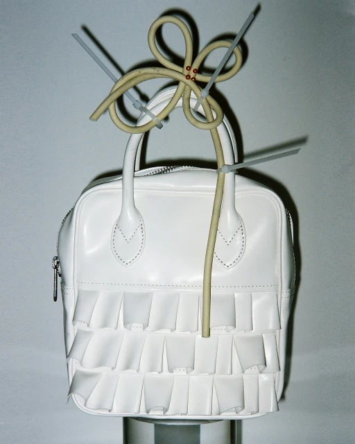 White purse on display, adorned with a flower made of electrical wire and held together with gray zip ties, on a gradient gray backdrop.