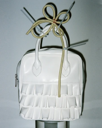 White purse on display, adorned with a flower made of electrical wire and held together with gray zip ties, on a gradient gray backdrop.