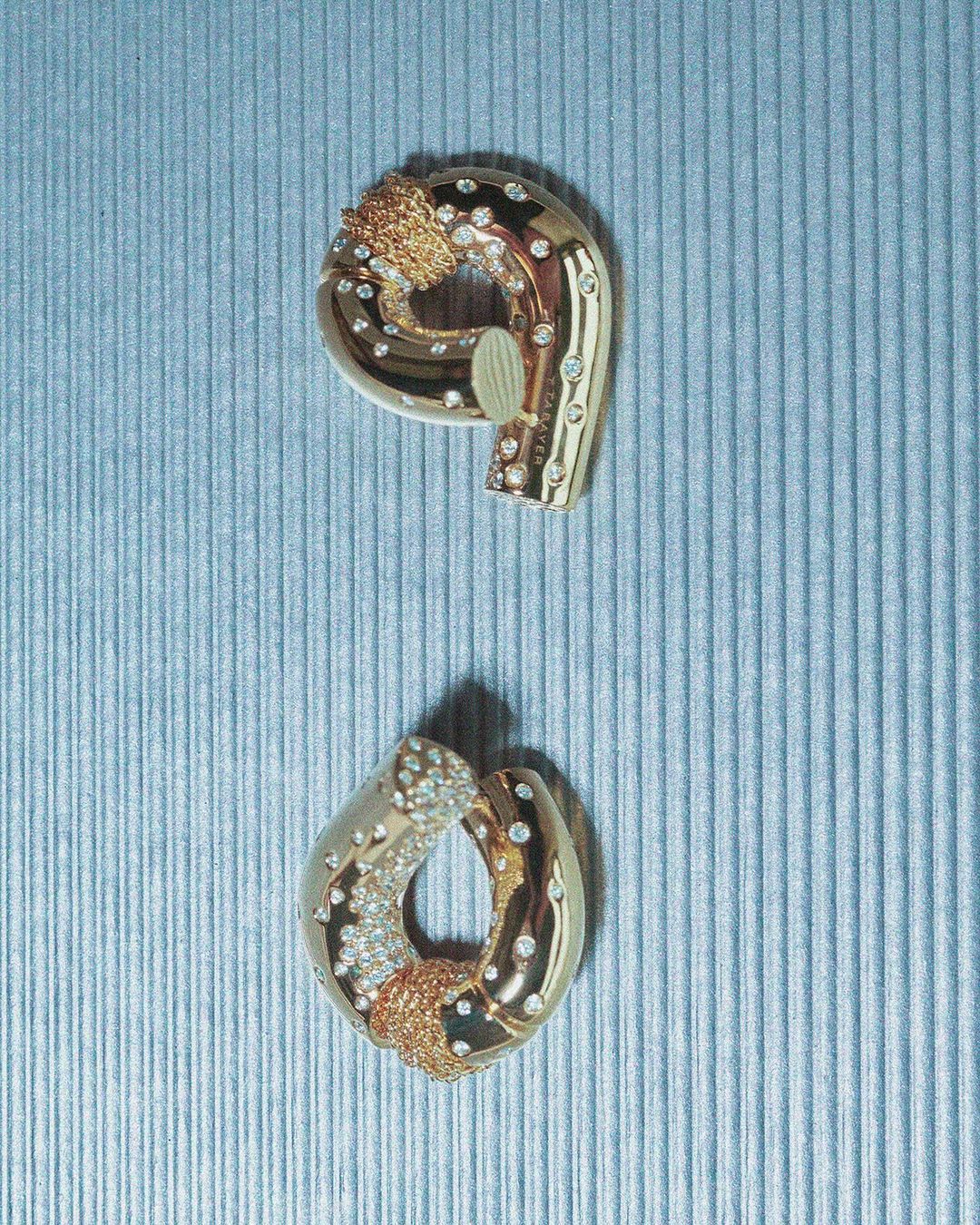 Two gold-toned jewellery pieces featuring gem stone accents are presented against a grooved blue background, in an indoor studio-like setting.