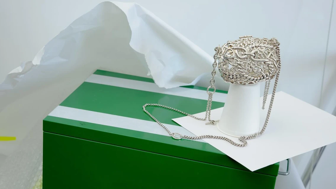 Silver-toned metal chainmail bag is displayed on a white paper cup, on a green and white striped surface, against a white backdrop.