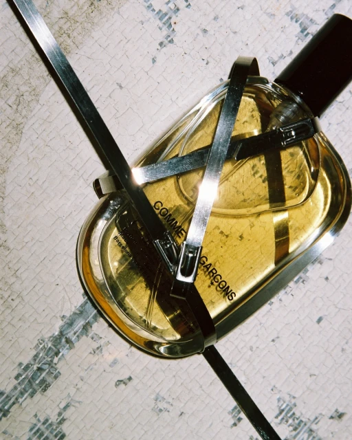 Close-up of a 'Comme des Garcons' perfume bottle with metal accents, set against a weathered, cracked surface with a subtle studio lighting.