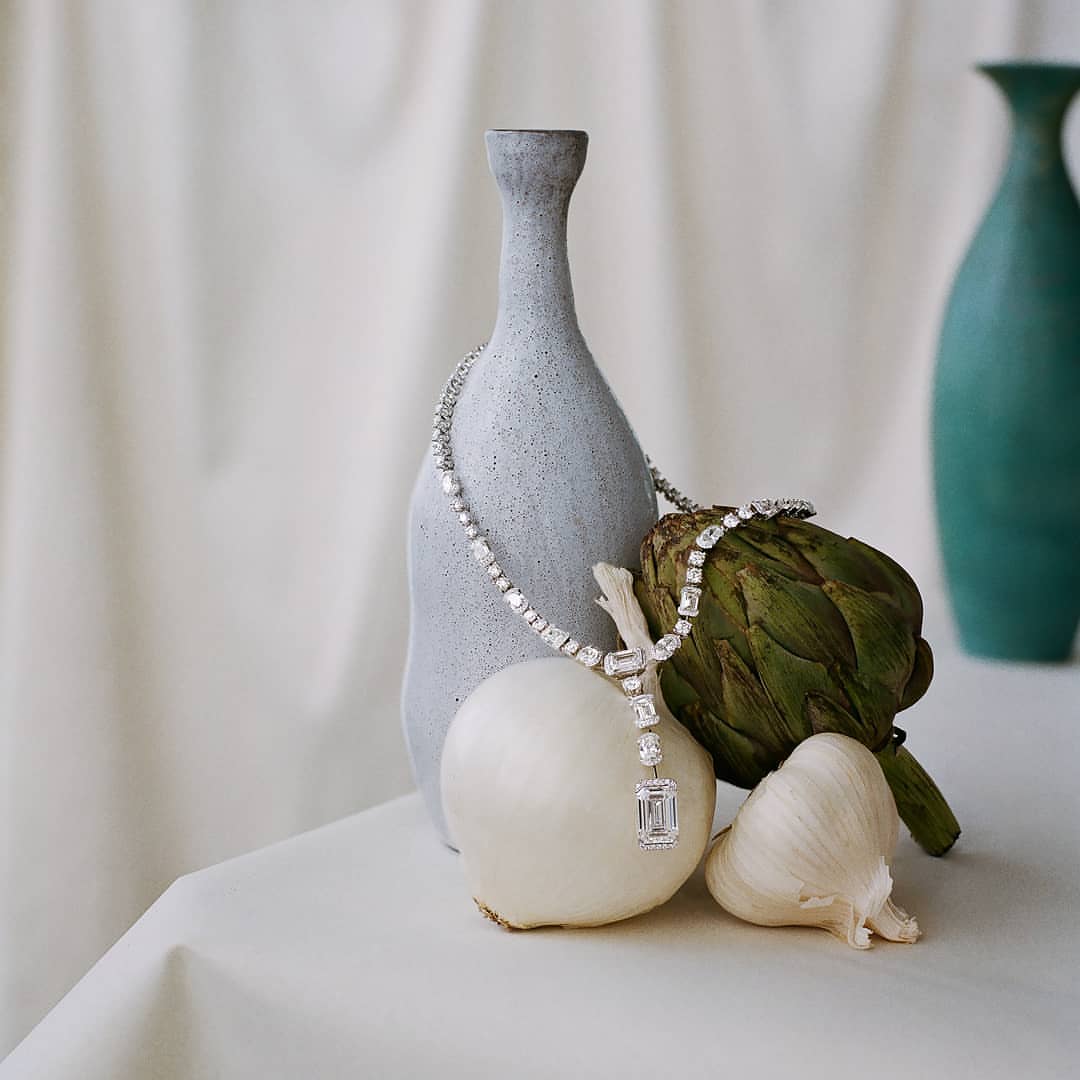 Still life shot of a diamond necklace draped over an artichoke and onions next to a gray vase on a white surface with a teal vase in background.