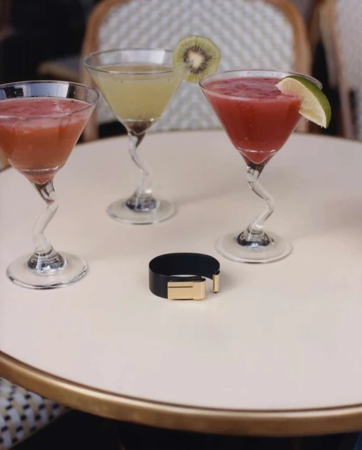 Three cocktail glasses filled with colorful drinks garnished with fruit, on a round white table next to a black and gold bracelet.