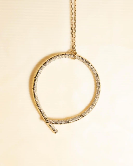 Elevated shot of a circular gold necklace featuring clear gems, gracefully suspended against a soft, neutral backdrop. Minimalist aesthetic.