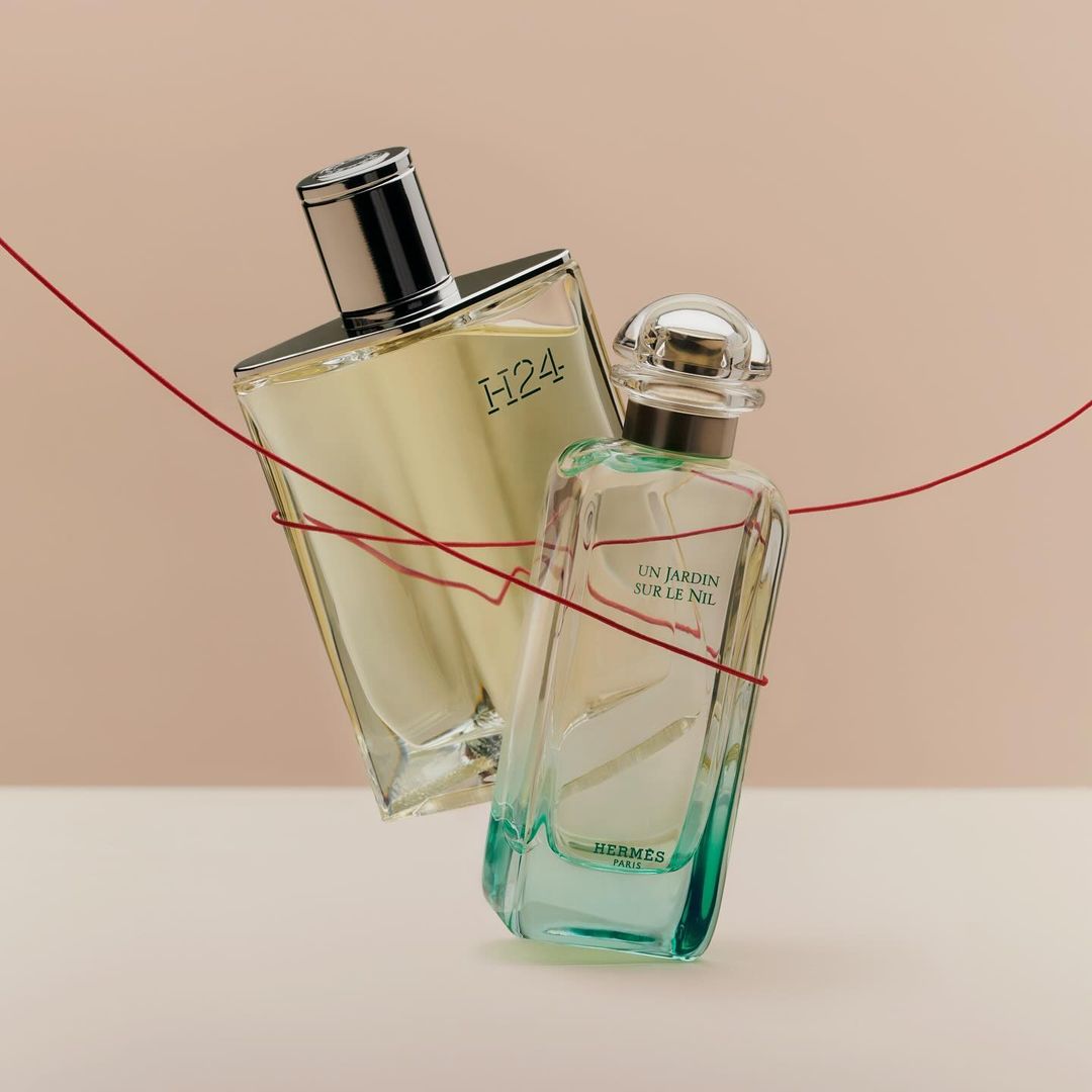 Two clear glass perfume bottles, one with a silver cap and the other with a clear cap, are connected by a red thread against a tan background.