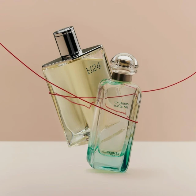 Two clear glass perfume bottles, one with a silver cap and the other with a clear cap, are connected by a red thread against a tan background.
