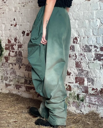 A person is wearing green, baggy pants and black boots, standing on straw in front of a distressed, white-painted brick wall.