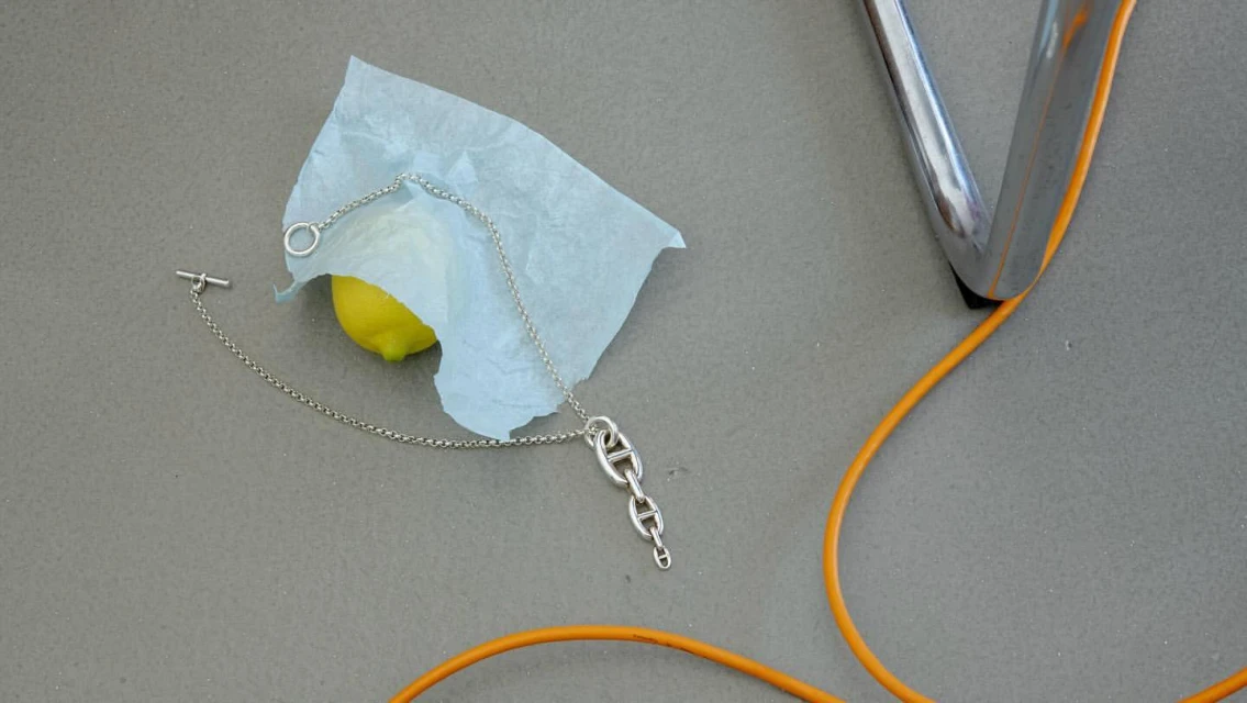 Silver chain necklace on a lemon wrapped in tissue paper, resting on a gray surface near a chrome chair leg with an orange cord.