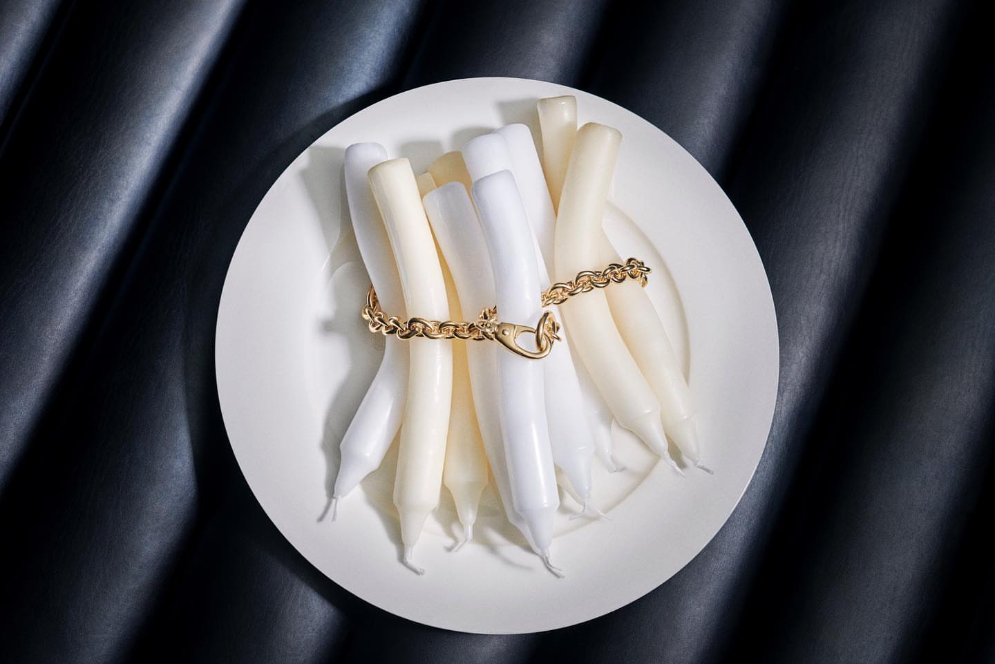 Elevated studio shot showcases a collection of white taper candles arranged on a white plate, fastened with a gold chain.