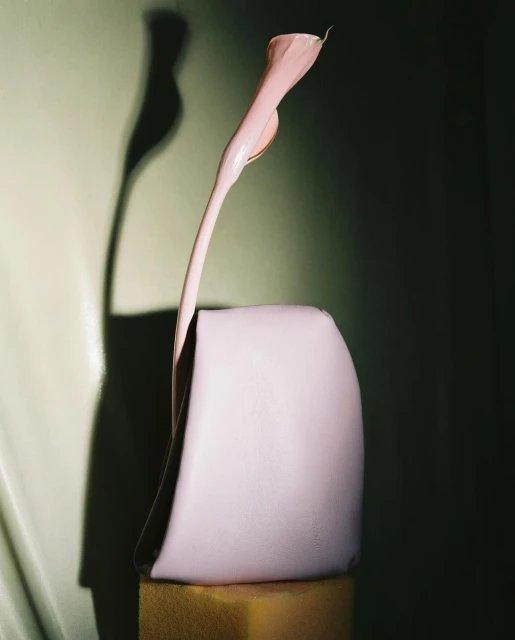 Still life studio photography of a pastel pink handbag propped on a gold block with a stylized calla lily and a dark green background.