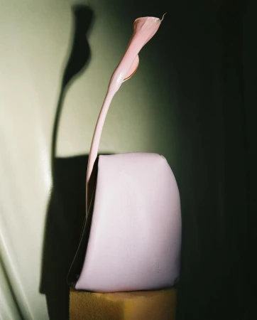 Still life studio photography of a pastel pink handbag propped on a gold block with a stylized calla lily and a dark green background.