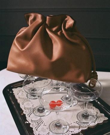 Tan leather pouch handbag resting on top of empty coupe glasses on a silver tray with a lace doily.