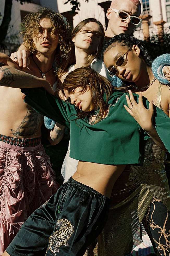 A diverse group of five young adult fashion models pose outdoors with natural light, showcasing unique streetwear styles, bold colors, and textures.