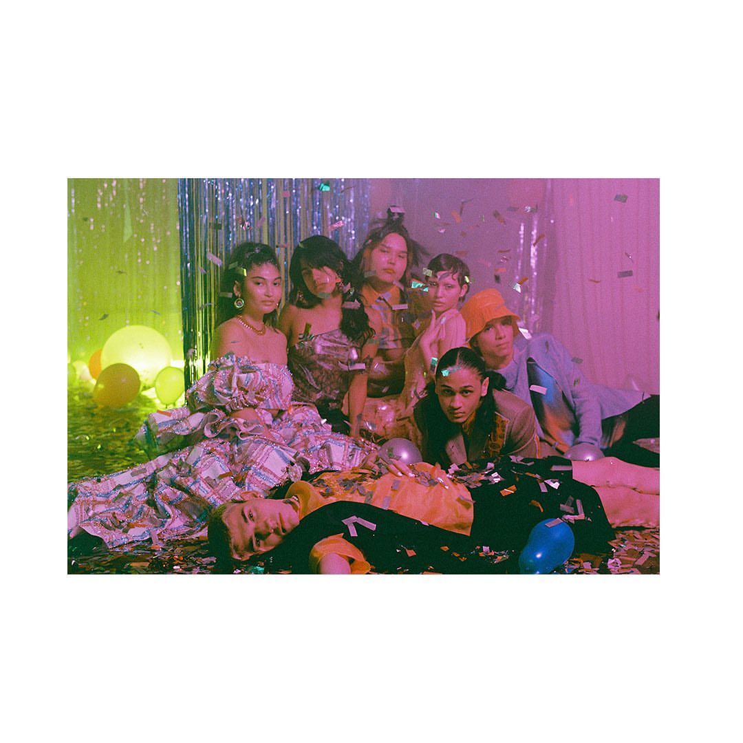 A group of seven diverse young adults in party attire pose among balloons and confetti in a studio environment with green and pink lighting.