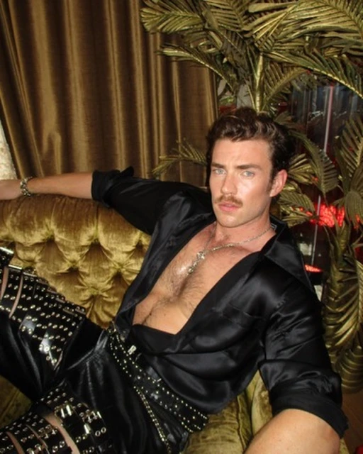 Man in black satin shirt and studded pants poses on gold velvet couch, indoor with potted palm tree.