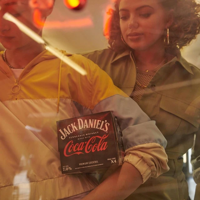 Two people stand near a display of Jack Daniel's mixed with Coca-Cola, with the beverage prominently centered in the frame.