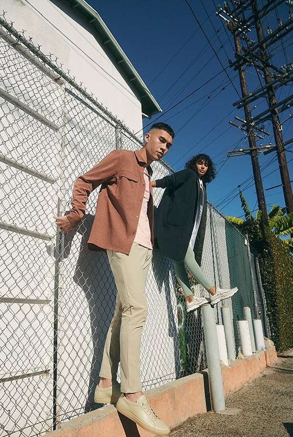 Two stylish adults, a male and female, pose on a concrete ledge in urban outdoor setting. The scene is framed by a chain-link fence and sky.