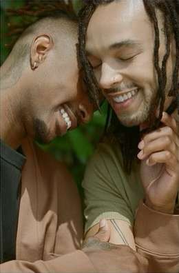 Two smiling African-American men with dreadlocks, wearing casual clothing, embrace closely, showing affection outdoors with green foliage.
