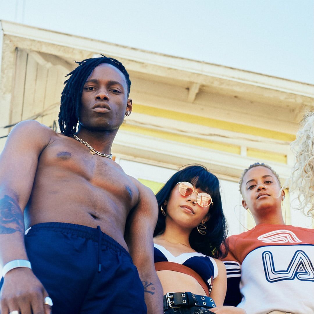Group of four young adult fashion models pose outside a white building with a shallow depth of field and natural light, captured at eye-level.
