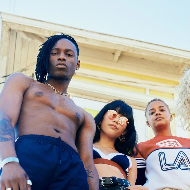 Group of four young adult fashion models pose outside a white building with a shallow depth of field and natural light, captured at eye-level.