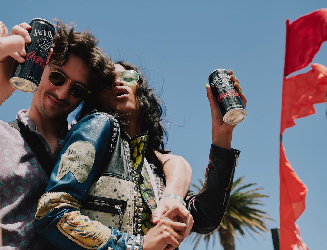 Two people hold Jack Daniel's and Coca-Cola canned cocktails outside on a sunny day with red flags and palm trees in the background.