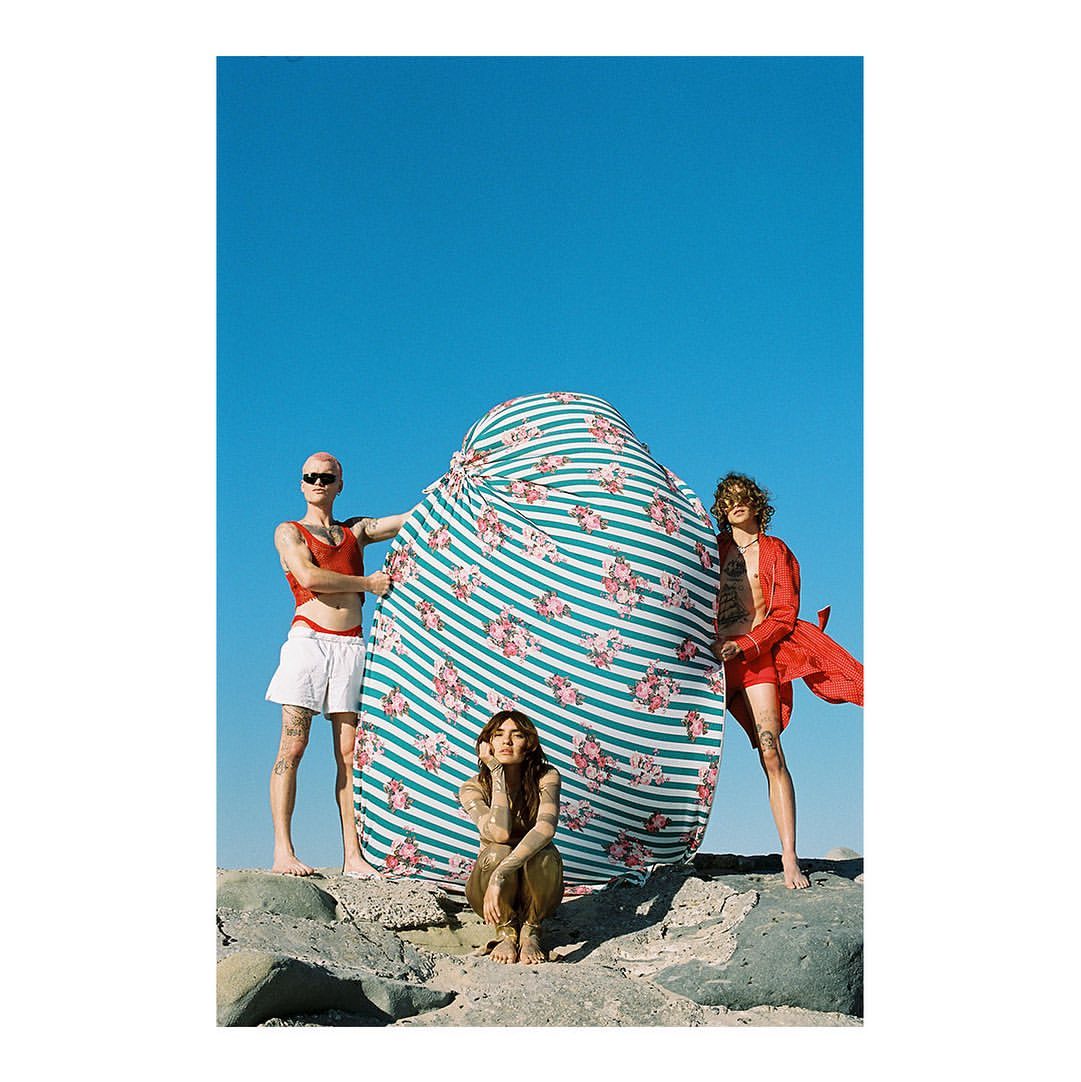 Three barefoot people with a striped and floral textile on rocky terrain under a clear blue sky, posing in an outdoor setting.