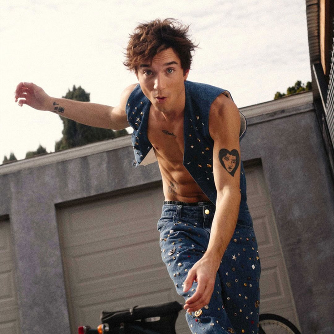Young adult male poses in embellished denim vest and jeans with tattoos, standing outdoors by a garage and scooter.