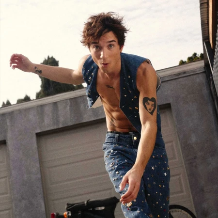Young adult male poses in embellished denim vest and jeans with tattoos, standing outdoors by a garage and scooter.